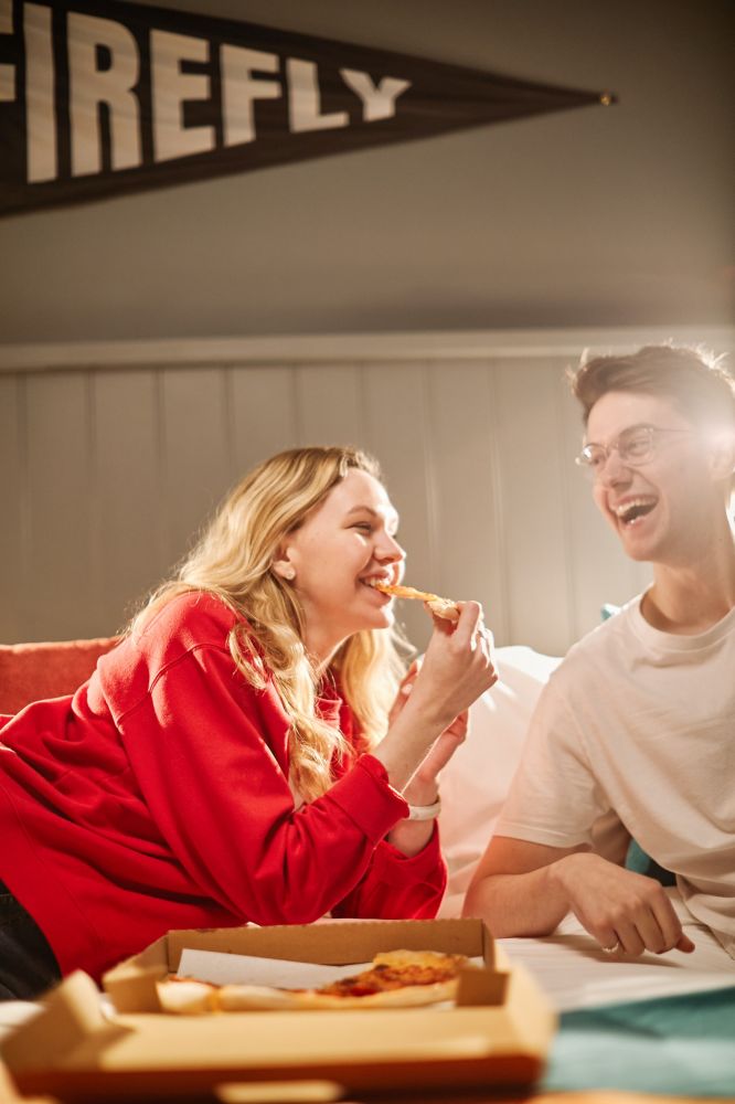 Hotel guests laying on double bed eating pizza